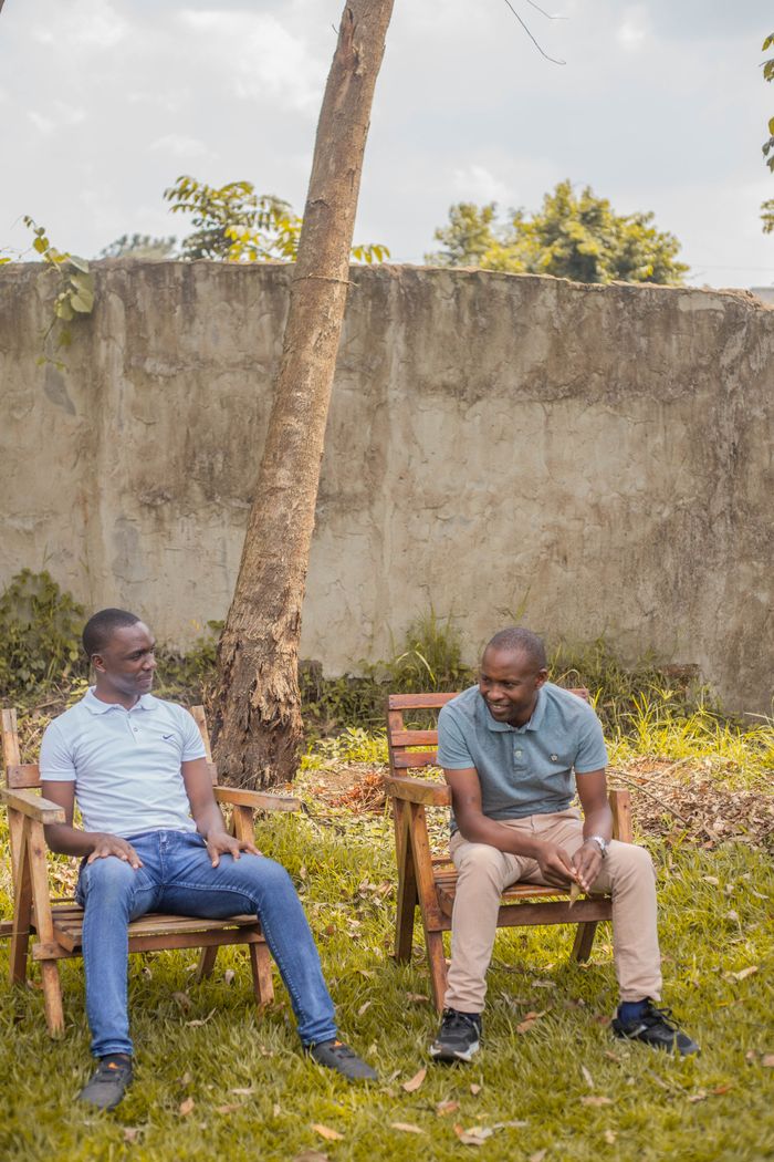 Two cheerful men sit talking on wooden chairs in a sunny outdoor garden setting.